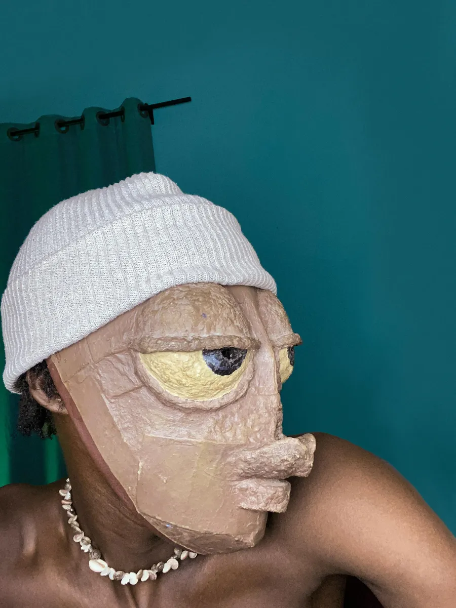 Edah wearing a handmade clay mask and a beanie, with a cowrie shell necklace, against a teal wall